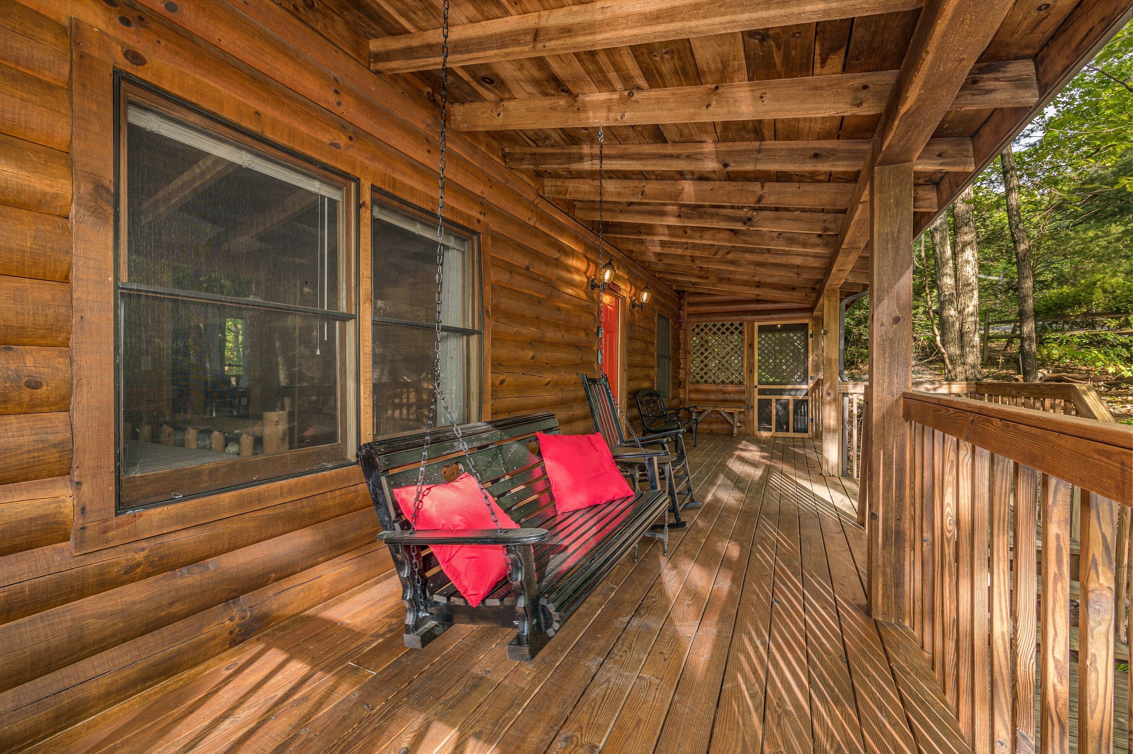 A wooden log cabin with a porch featuring a bench with red cushions, large windows, and a view of the surrounding forest.