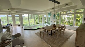 A spacious room with a large dining table, multiple chairs, and a sofa. There are large windows providing a view of lush greenery outside. - Spacious Tropical Oasis (Princeville)