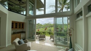 A spacious living area with a large sliding glass door leading to a patio with outdoor furniture and a lush garden view. - Spacious Tropical Oasis (Princeville)