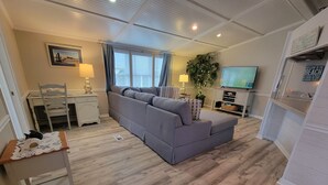 A living room with a grey sofa, a TV, a plant, and a desk with a chair. - **NEW FLOORS** BEACH HOUSE, CENTRAL AIR, GOLF CART INCLUDED (Myrtle Beach)