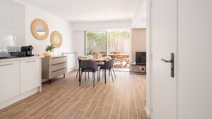 A modern kitchen with white cabinets, a coffee machine, and a dining area with a table and chairs. - 2-Bedroom Apartment with Garden (Cannes)