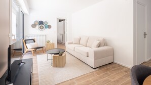 A modern living room with a sofa, a coffee table, and a TV. There is a window, a radiator, and a door in the background. - 2-Bedroom Apartment with Garden (Cannes)