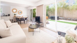 A modern living room with a sofa, a coffee table, and a TV. There is a dining area with a table and chairs. The room has large windows with a view of a garden and a building in the background. - 2-Bedroom Apartment with Garden (Cannes)