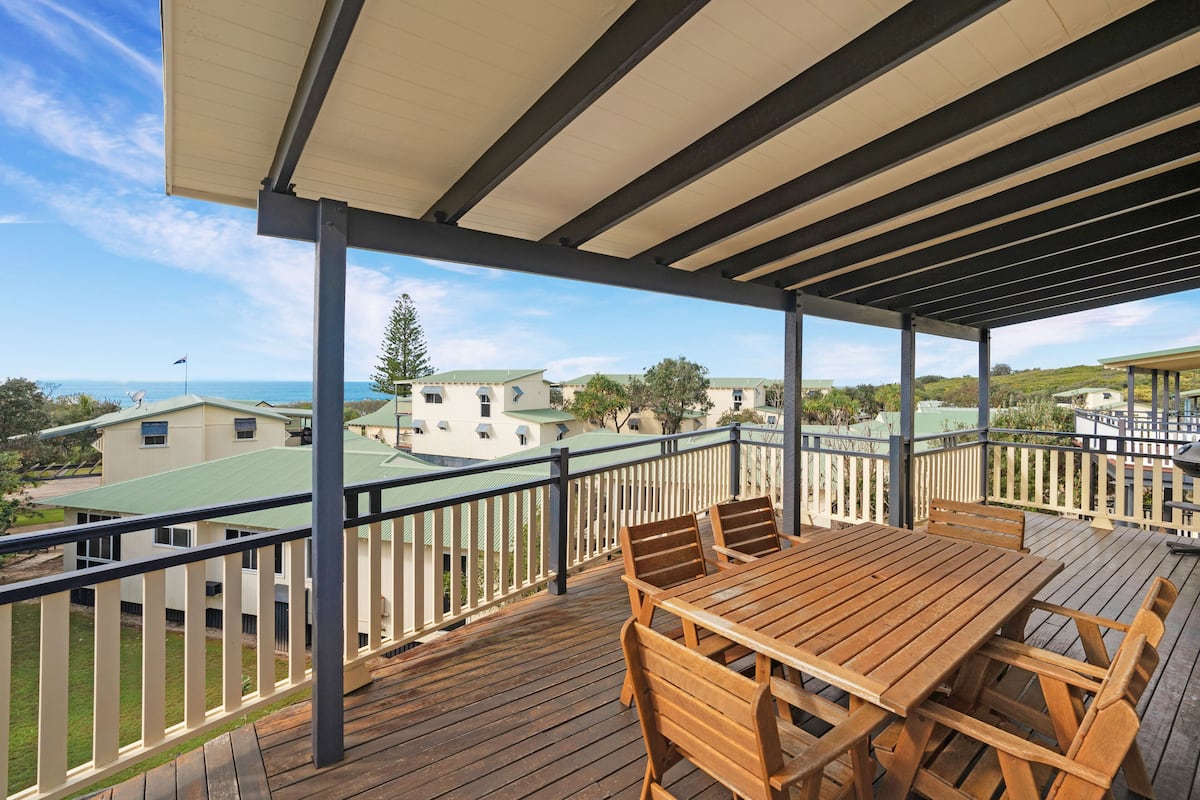 A wooden deck with a table and chairs, overlooking a residential area with houses and a view of the ocean.