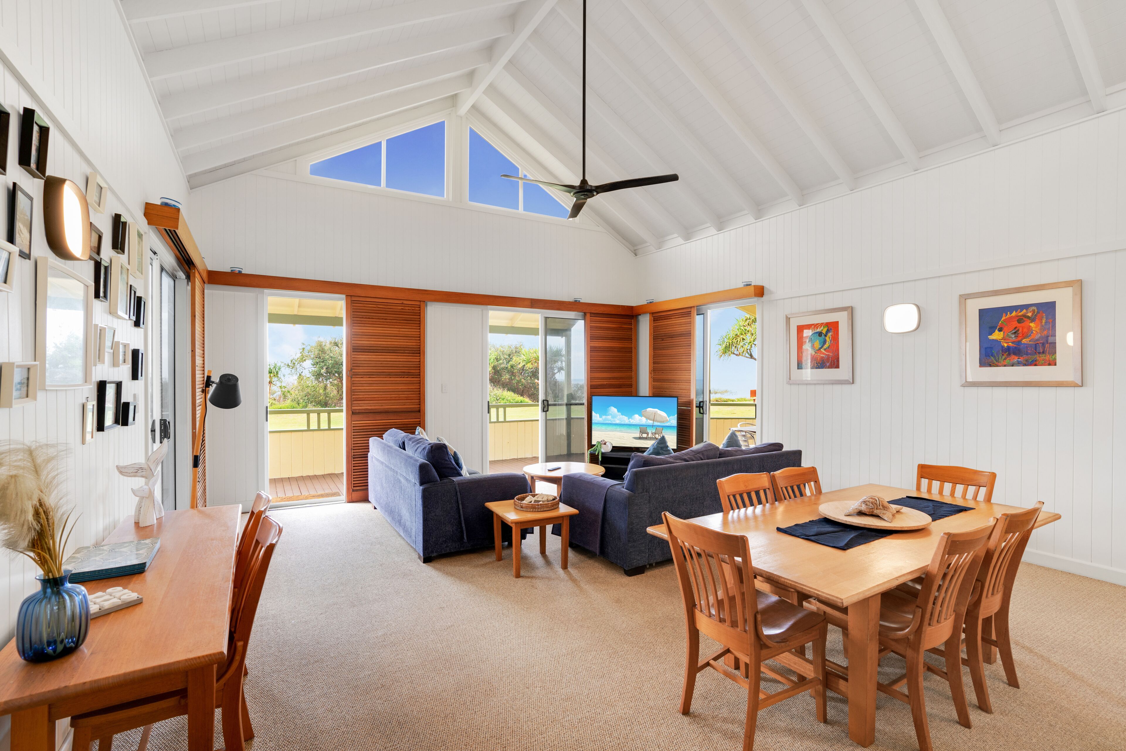 A spacious living area with a large sofa, dining table, and chairs. There is a television on a stand, a ceiling fan, and a view of the outdoors through large windows.