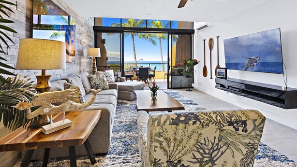 Een moderne woonkamer met een grote televisie, comfortabele zithoek, een plafondventilator en uitzicht op de oceaan door grote ramen.