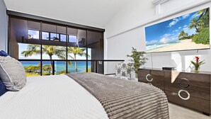 Een slaapkamer met een groot bed, een aan de muur bevestigde televisie en uitzicht op het strand door middel van ramen van de vloer tot aan het plafond.