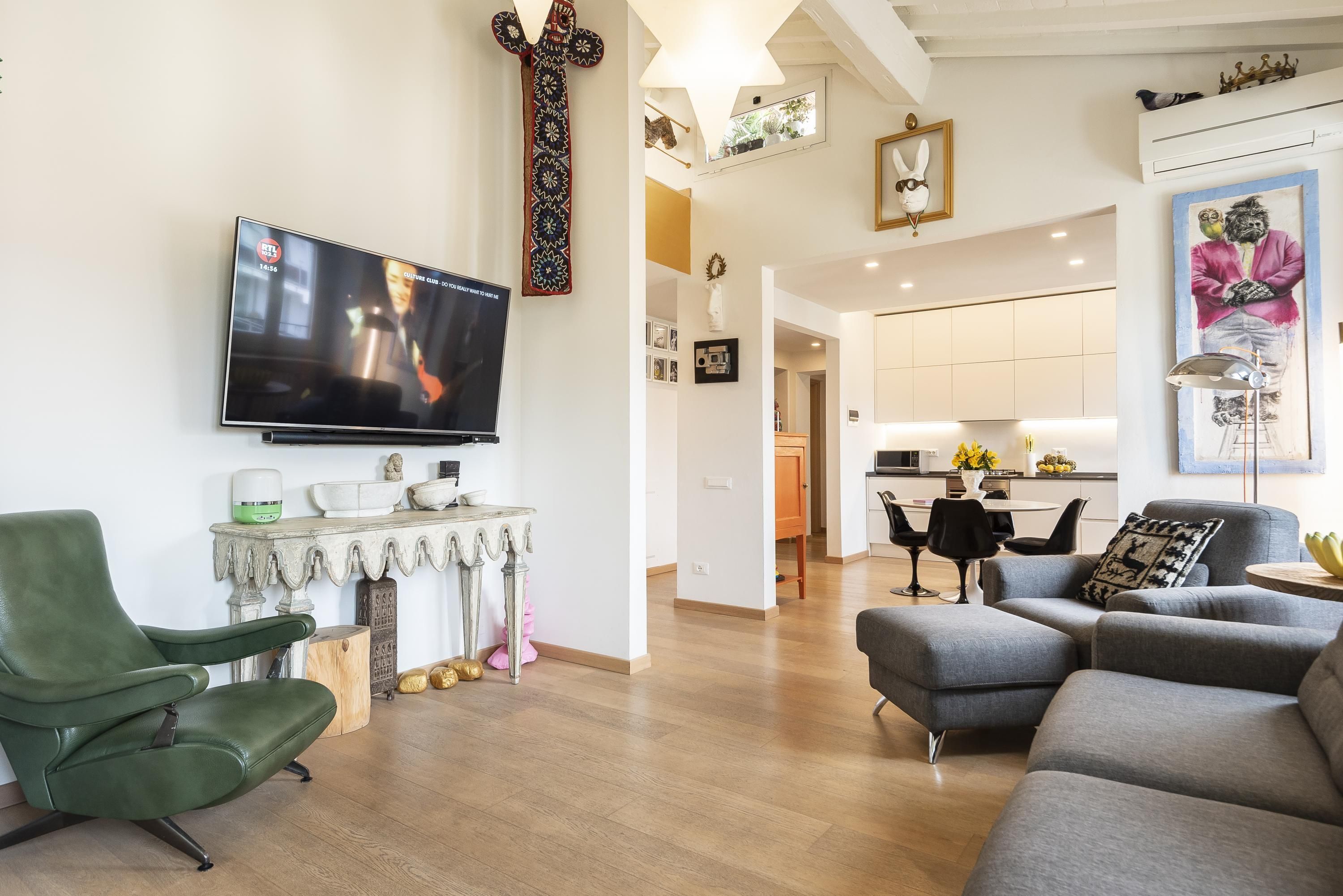 A modern living room with a flat-screen TV, a green armchair, a grey sofa, and a kitchen in the background.