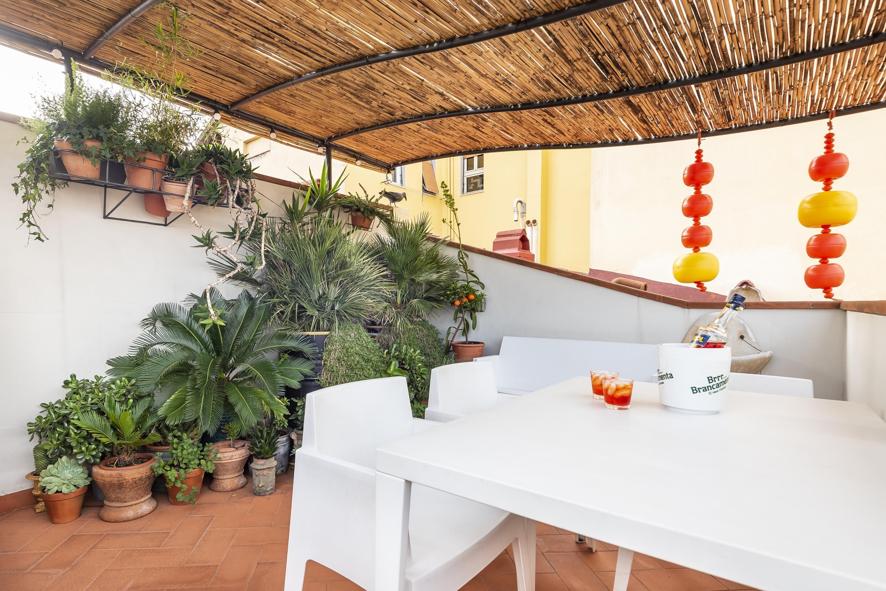 A covered patio area with a white table, potted plants, and a bottle of Branca Branca wine.