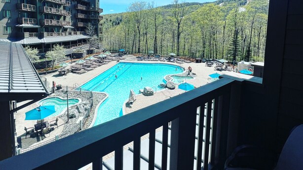 A resort with a large swimming pool, outdoor seating, and a view of a forested mountain. - Spruce Peak, Studio -Ski in -Ski out (Stowe)