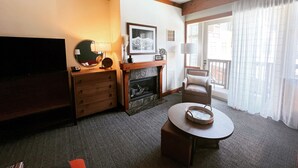 A hotel room with a flat-screen TV, a fireplace, a round table with a basket, a sofa, and a large window with curtains. - Spruce Peak, Studio -Ski in -Ski out (Stowe)