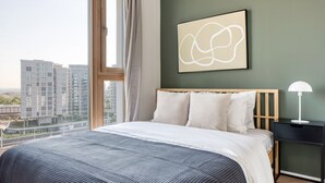 Ein modernes Hotelzimmer mit einem großen Bett, einem Nachttisch, einer Lampe und Blick auf hohe Gebäude.