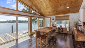 A spacious dining area with a wooden table and chairs, large windows offering lake views, and a wooden ceiling. - A Point to Happiness by Lake Martin Vibe (Dadeville)