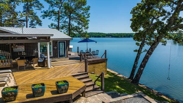 A wooden deck with outdoor seating, a blue umbrella, and a view of a lake surrounded by trees. - A Point to Happiness by Lake Martin Vibe (Dadeville)