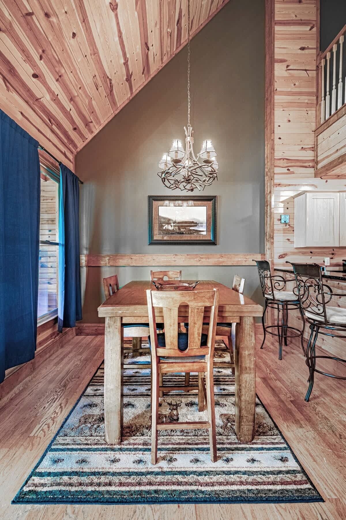 A dining area with a wooden table and chairs, a chandelier, a framed picture on the wall, and a view of the outdoors through a window.