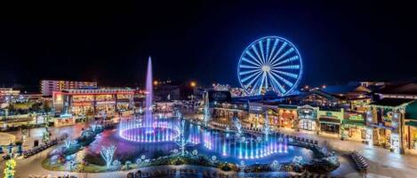 A nighttime view of a city with a large illuminated Ferris wheel, a fountain, and various buildings.