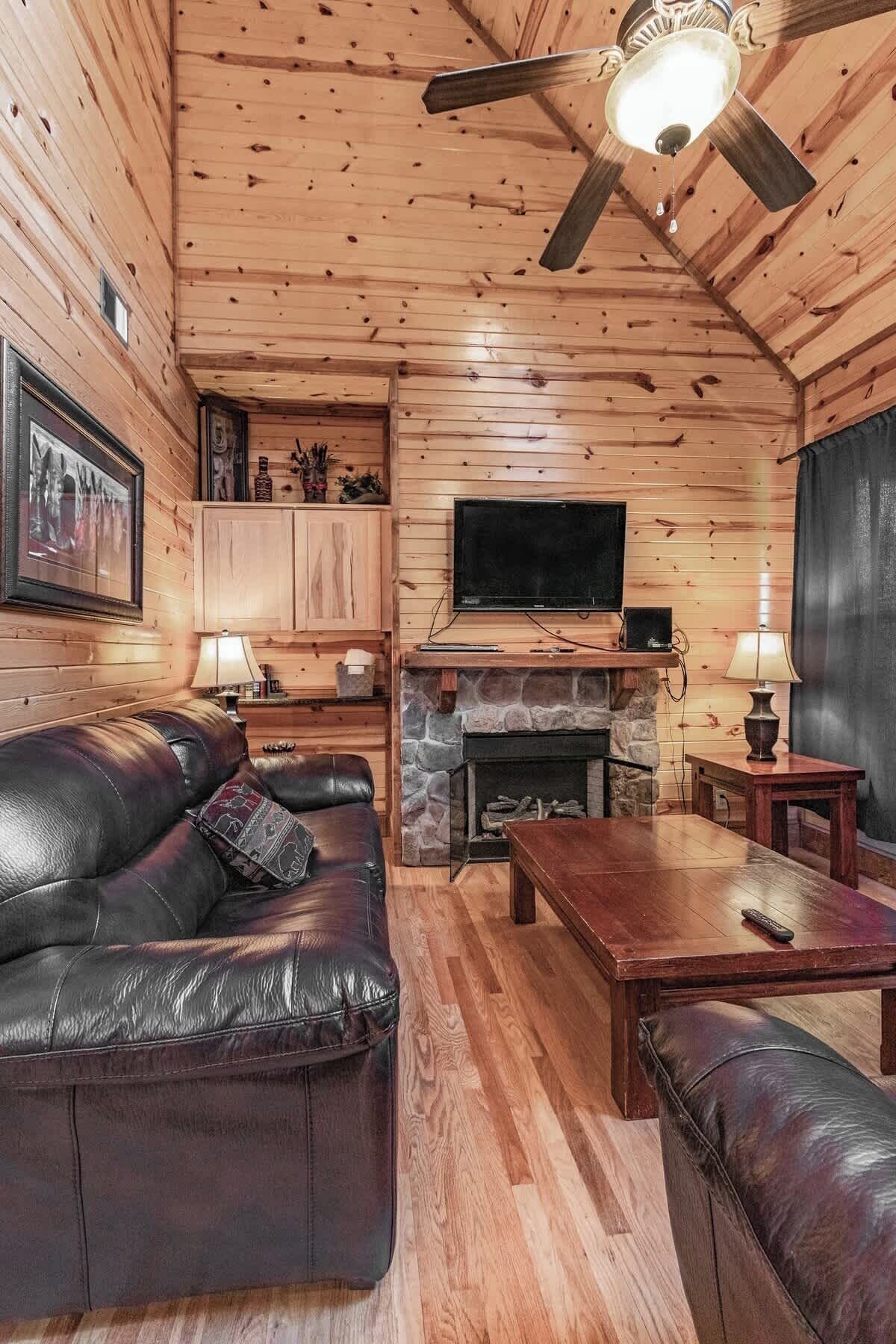 A log cabin interior with a leather sofa, a stone fireplace, a television, and a ceiling fan.