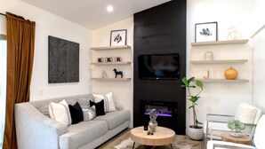 A modern living room with a grey sofa, a round coffee table, and a fireplace. There are shelves with decorative items and a TV mounted on the wall.