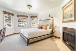 Quarto com uma cama grande, três janelas, lareira e mesa de cabeceira com abajur.