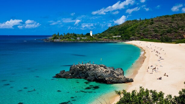 A beach with clear turquoise waters, a rocky outcrop, and a white lighthouse on a hill. - Spacious North Shore cottage with lush mountain scenery in the country! (Waialua)
