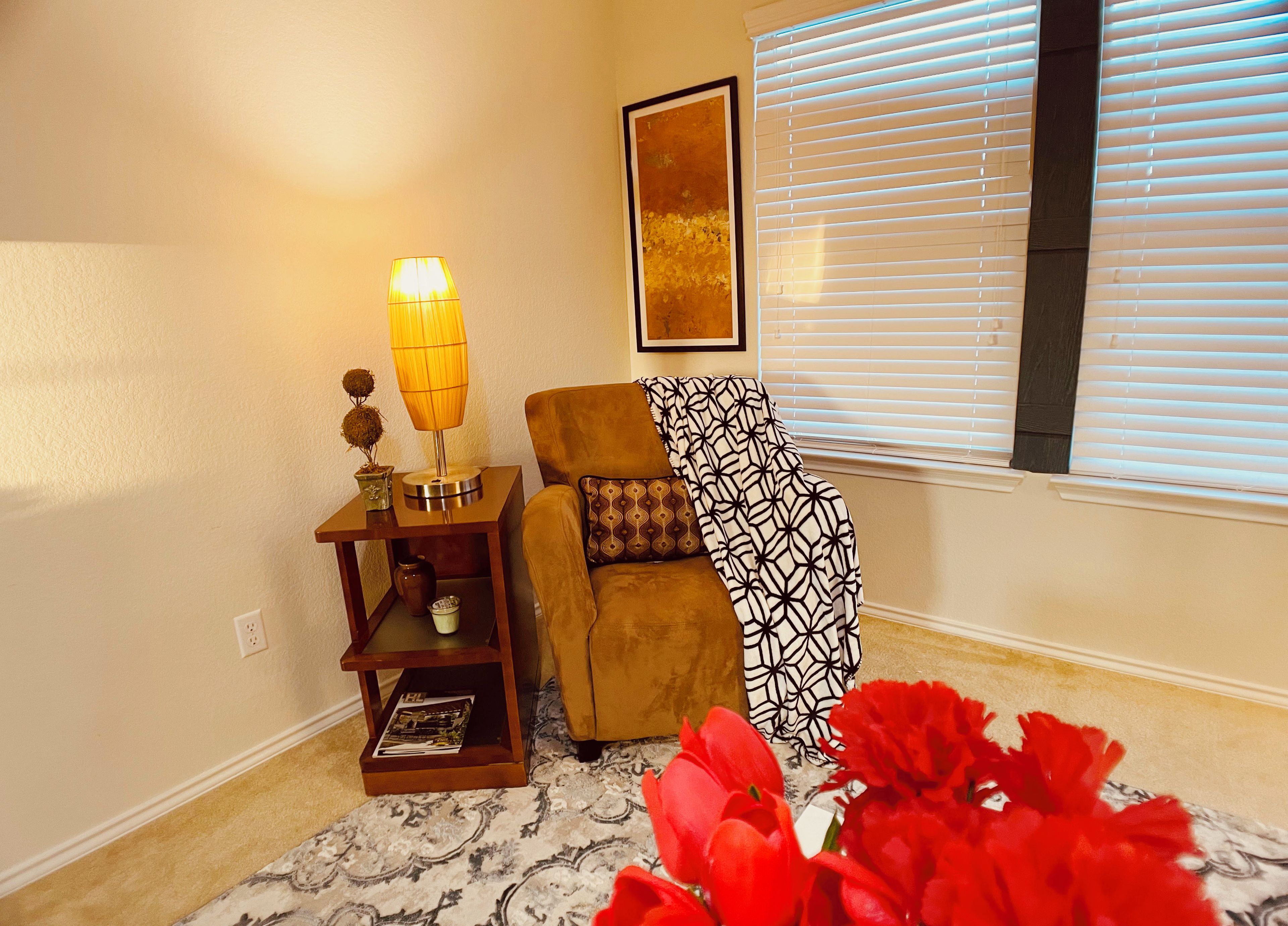 A cozy living room with a brown sofa, a patterned throw, a lamp, and a vase with red flowers.