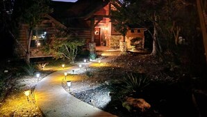 Una cabaña de troncos con fachada de piedra, un camino de hormigón iluminado por luces del suelo y un jardín cuidado.