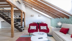 Un loft moderno con vigas de madera a la vista, una escalera, un sofá, dos sillones rojos y una pequeña mesa con un control remoto.