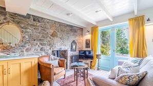 A cozy living room with a stone wall, a fireplace, a television, and a large window with curtains. - Secret Cottage, St Ives – Sleeps 6 – Sea Views – Garden (St Ives)