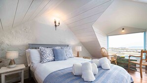 A bedroom with a bed, two bedside tables, a lamp, a chair, and a view of the sea. - Secret Cottage, St Ives – Sleeps 6 – Sea Views – Garden (St Ives)