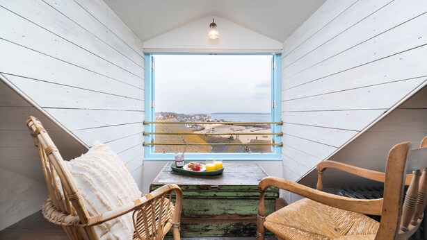 A balcony with a view of the city, two wicker chairs, a table with a plate of food and drinks, and a hanging light fixture. - Secret Cottage, St Ives – Sleeps 6 – Sea Views – Garden (St Ives)