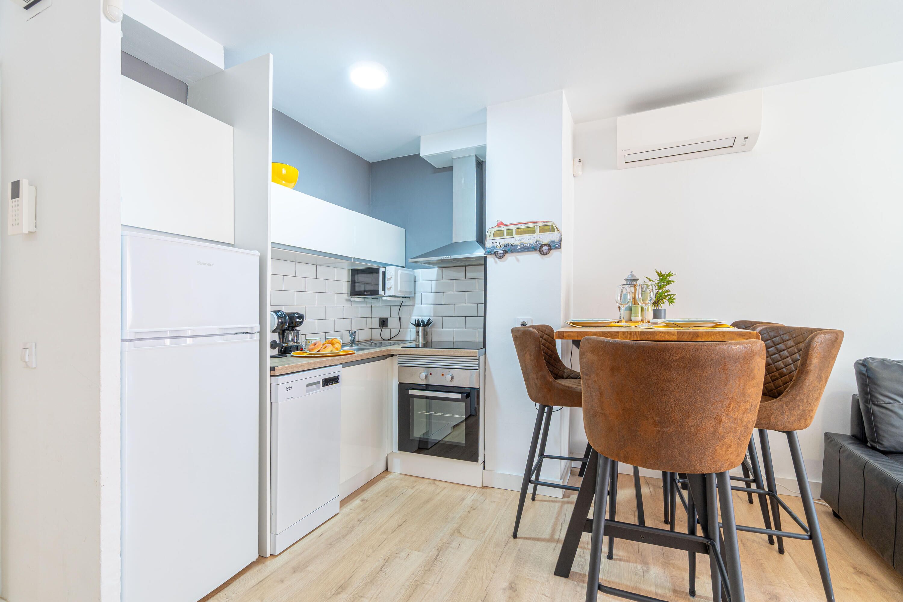 A modern kitchen with white cabinets, a built-in oven, a microwave, and a coffee machine. There is a dining area with a table and two chairs.