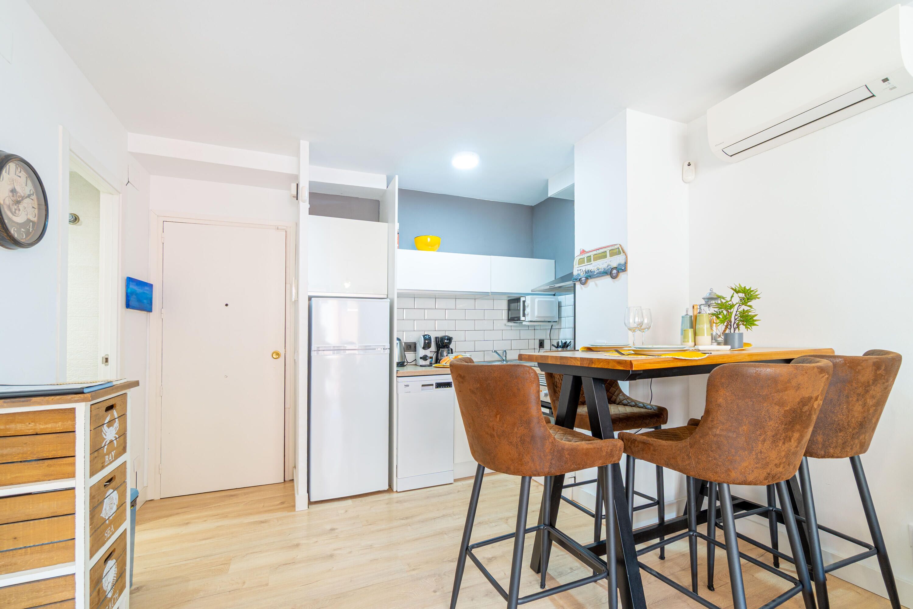 A compact kitchen with a white refrigerator, a microwave, and a coffee machine. There is a dining table with four brown chairs and a small potted plant on it.
