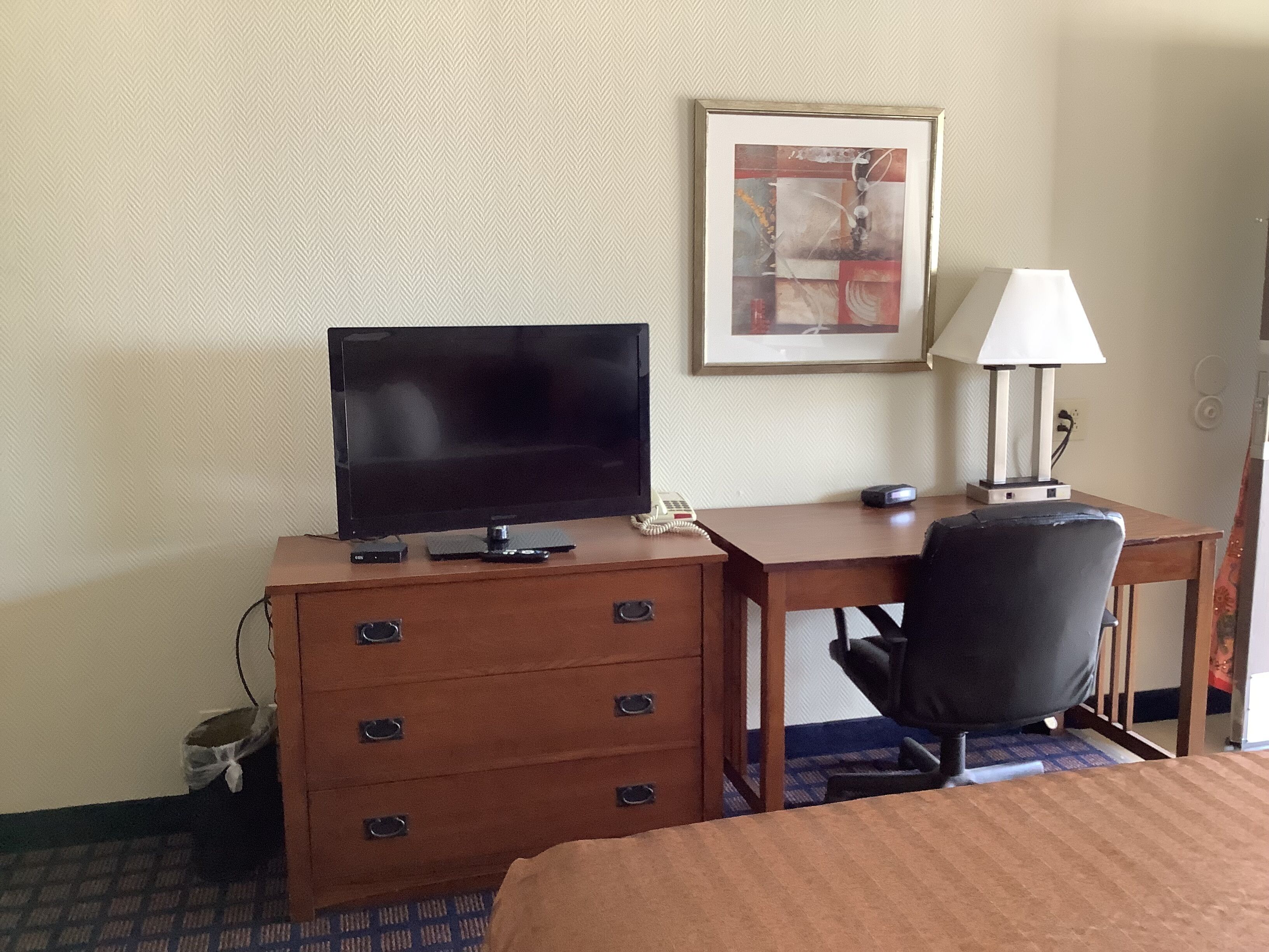 A hotel room with a bed, a desk with a flat-screen TV, a chair, a desk lamp, and a framed abstract painting on the wall.