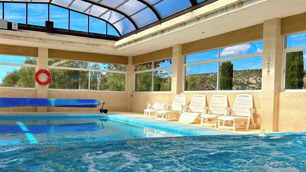 En inomhuspool med kristallklart blått vatten, ett glastak och vita solstolar längs poolkanten.