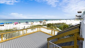Eine Holzterrasse mit Geländer, die auf einen Strand mit Sonnenschirmen und Liegestühlen blickt.