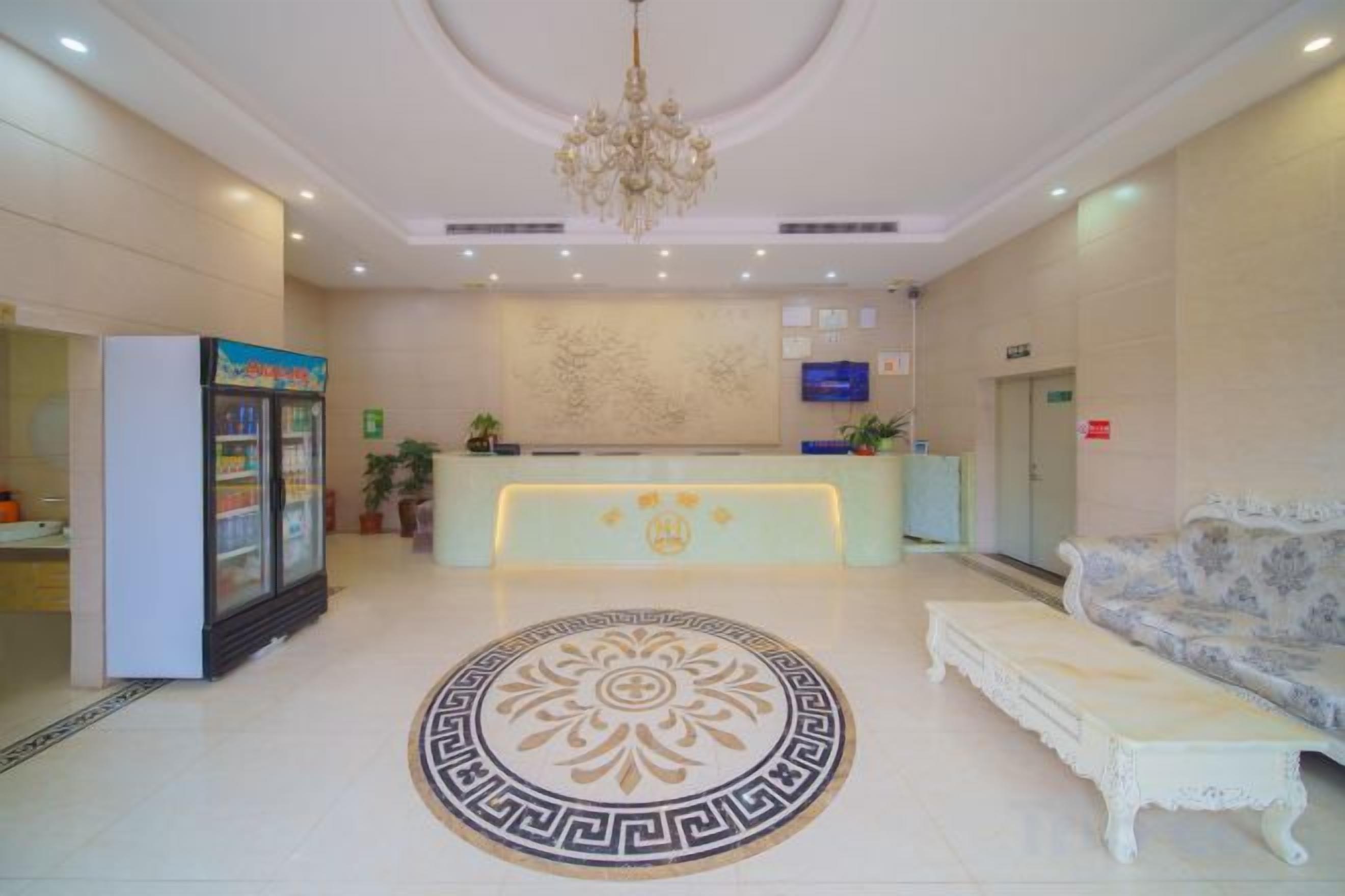 Un vestíbulo de hotel con una mesa de recepción, un candelabro, un suelo estampado, un sofá y un refrigerador.