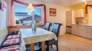 Een zithoek met een tafel, stoelen en een bank. Er is een raam met gordijnen, een hangende lamp en een keuken met kastjes en een koelkast.