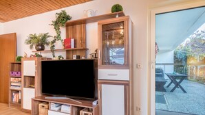 Een woonkamer met een televisie, houten kasten, plantenbakken en een schuifdeur naar een terras.