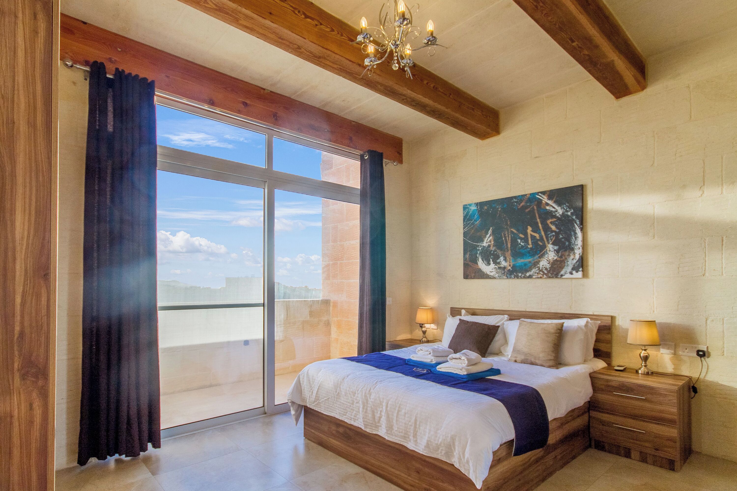 A bedroom with a large bed, wooden headboard, two bedside tables with lamps, a painting on the wall, and a sliding glass door leading to a balcony.