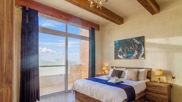 A bedroom with a large bed, wooden headboard, two bedside tables with lamps, a painting on the wall, and a sliding glass door leading to a balcony.