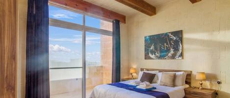 A bedroom with a large bed, wooden headboard, two bedside tables with lamps, a painting on the wall, and a sliding glass door leading to a balcony.