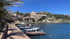 A marina with several boats docked, a sandy walkway, and buildings with Mediterranean architecture on a hillside. - Grecalotto Apartment (La Maddalena)