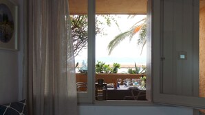 A room with a view of the beach, a dining area with a table and chairs, and a window with curtains. - Grecalotto Apartment (La Maddalena)