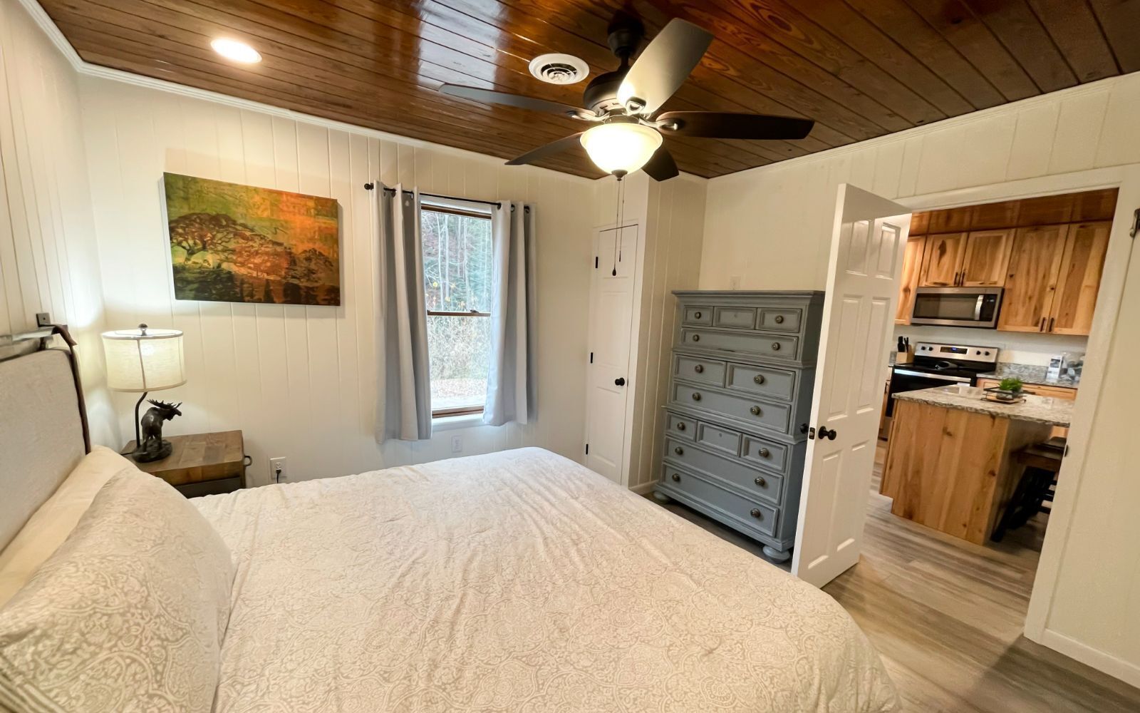 Un dormitorio con cama, mesita de noche, lámpara, ventilador de techo, ventana con cortinas y vista hacia una zona de cocina.