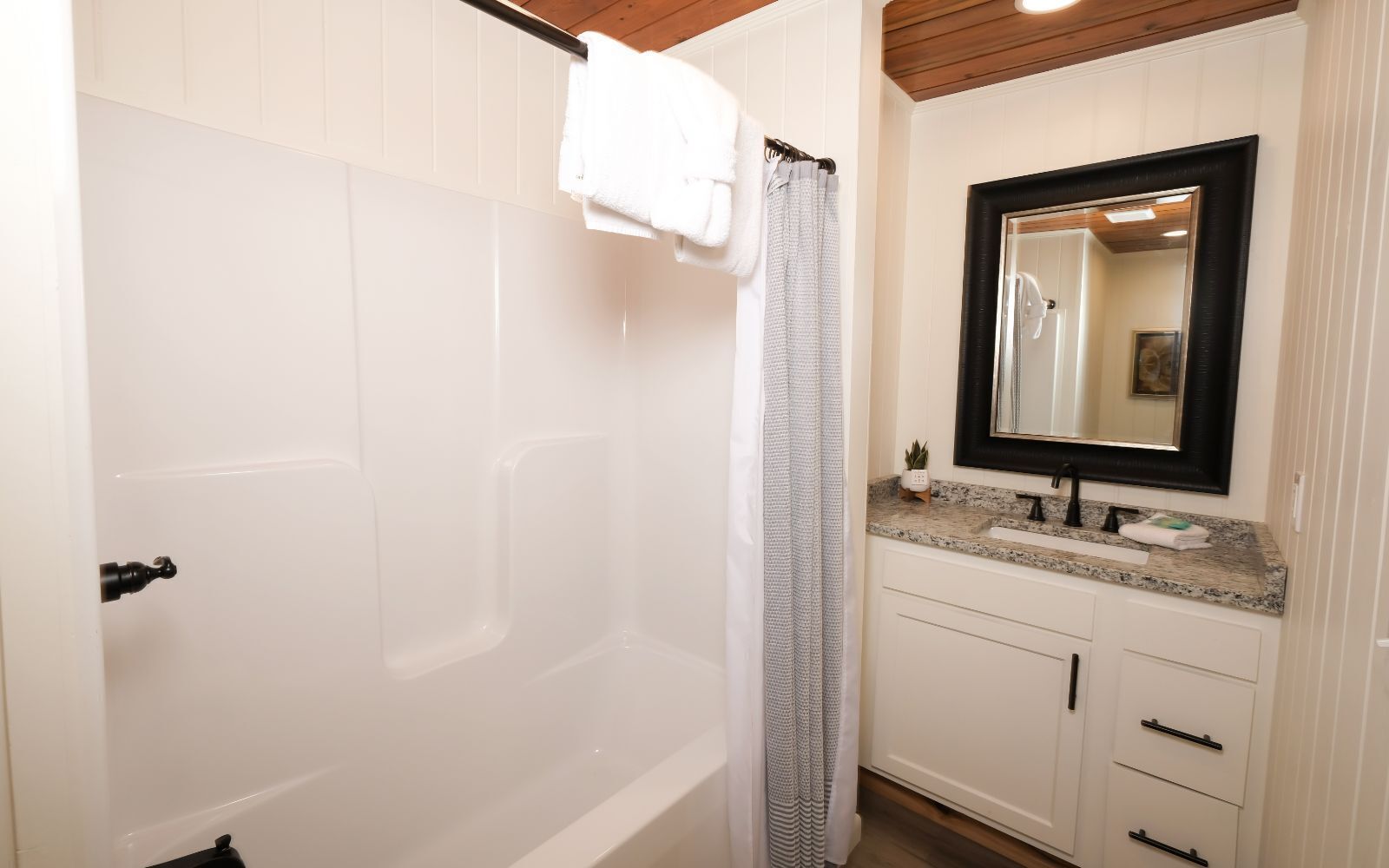 A bathroom with a white bathtub, a shower curtain, a sink with a granite countertop, and a mirror with a black frame.