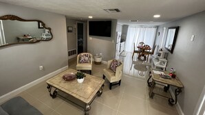 Un salon comprenant une table basse, deux fauteuils, une table à manger avec des chaises, un coin cuisine et un grand miroir.