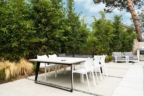 Un patio exterior con una mesa y sillas blancas, rodeado de abundante vegetación y un árbol.