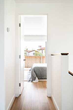 Un pasillo estrecho que conduce a un dormitorio con un amplio ventanal, suelo de madera y un área exterior visible con valla y edificios.