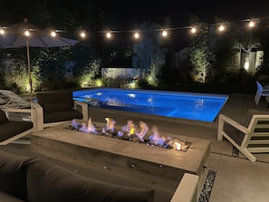 Un espace extérieur avec piscine, agrémenté d’un foyer intégré, de chaises longues et de guirlandes lumineuses.