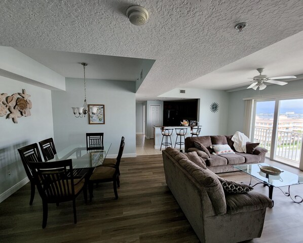 A spacious living area with a dining table, chairs, a sofa, and a balcony with a view. - Spacious, kid-friendly 3BR beachfront condo, close to Orlando theme parks (Daytona Beach)